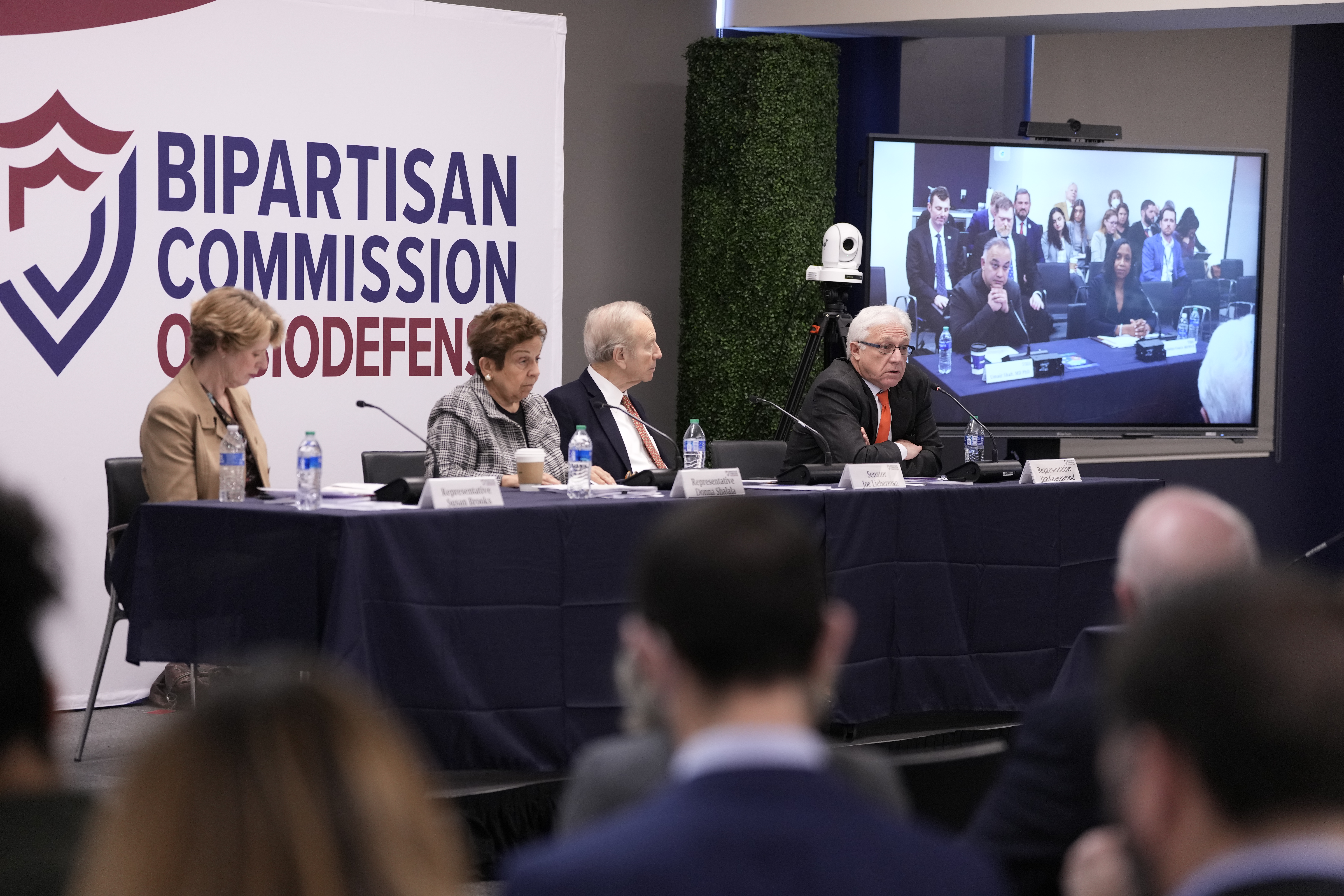 Four commission members sitting at a table in the front of the room with Dr. Shah on the screen behind them.