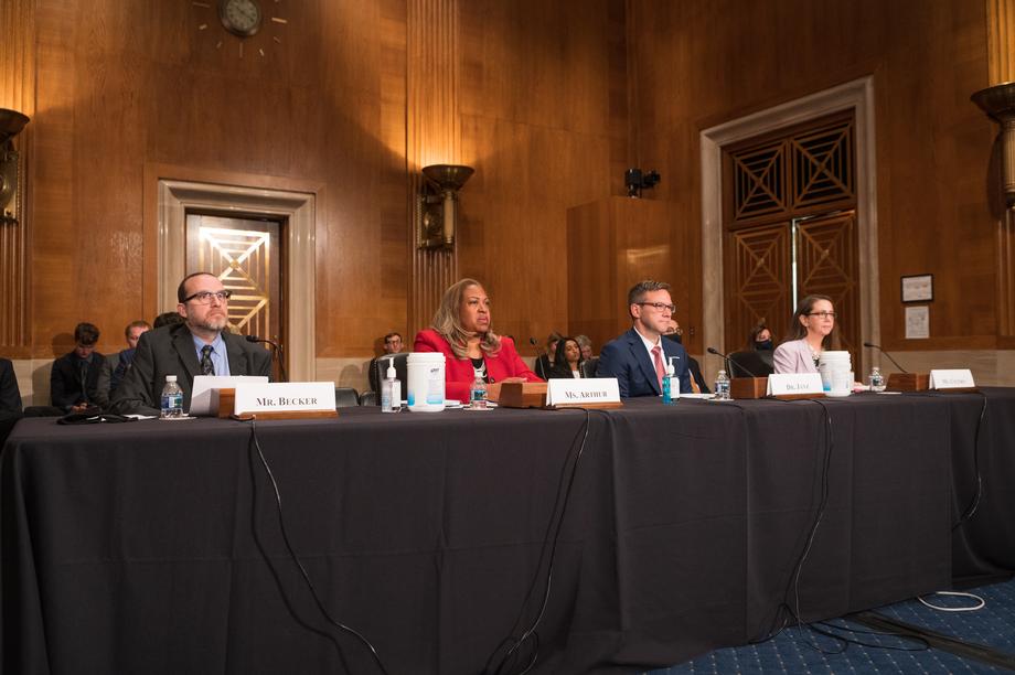 Four individuals giving their testimonies while sitting at a table with black tablecloth, name plates, microphones, and more.