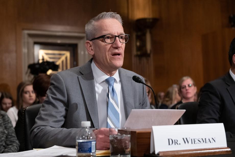 Dr. Wiesman, a gray haired man wearing glasses and gray suit, speaking into a microphone while seated at a table.