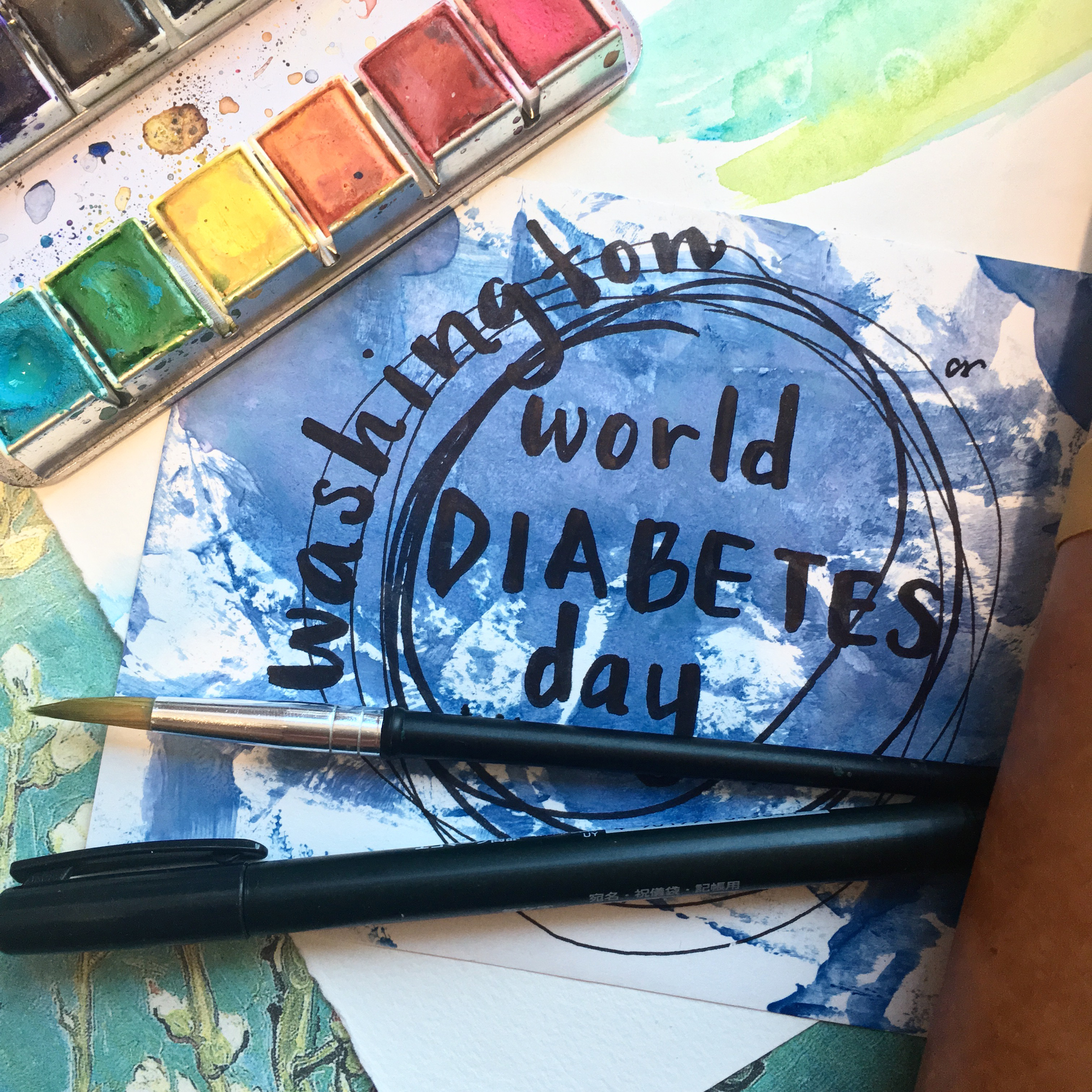 A piece of paper with blue paint with fancy lettering that says the words "Washington Diabetes Day" on top of the paining is a rainbow paint palate, a black paint brush, and a black felt tip pen.