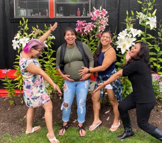 A group of 4 Pacific Islander women standing outside in front of lilies, smiling and pointing to their relative's pregnant stomach.