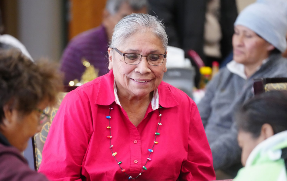 An older Tribal woman in a bright pink shirt smiles while seated at a table with a group.