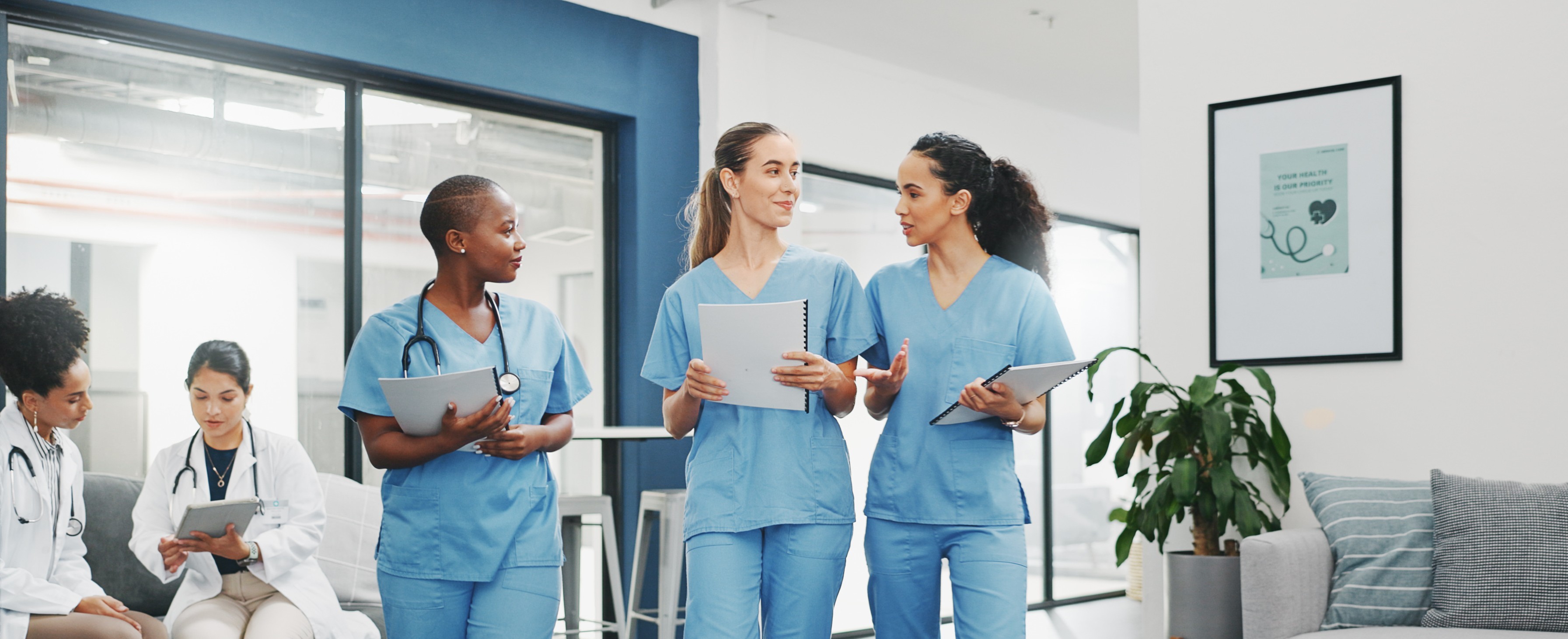 Three young women wearing blue medical scrubs and holding papers walk through an office lounge with large windows.