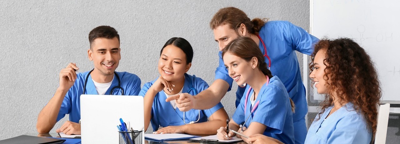 Two male and three female nursing students in blue scrubs smile looking at a laptop seated at a conference table.