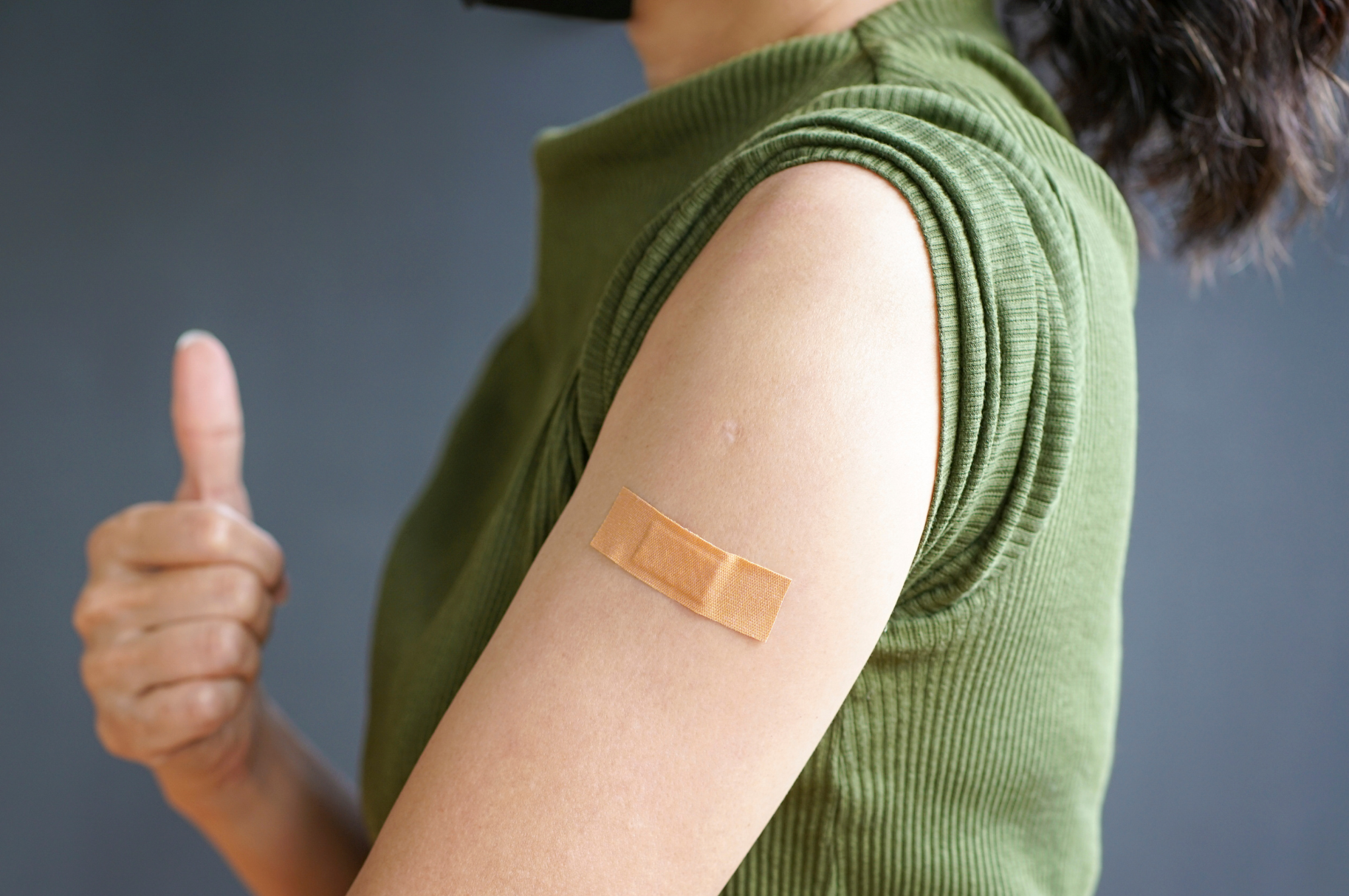 Close up of left arm of a white female showing a band-aid over a vaccination with right hand showing thumbs up..