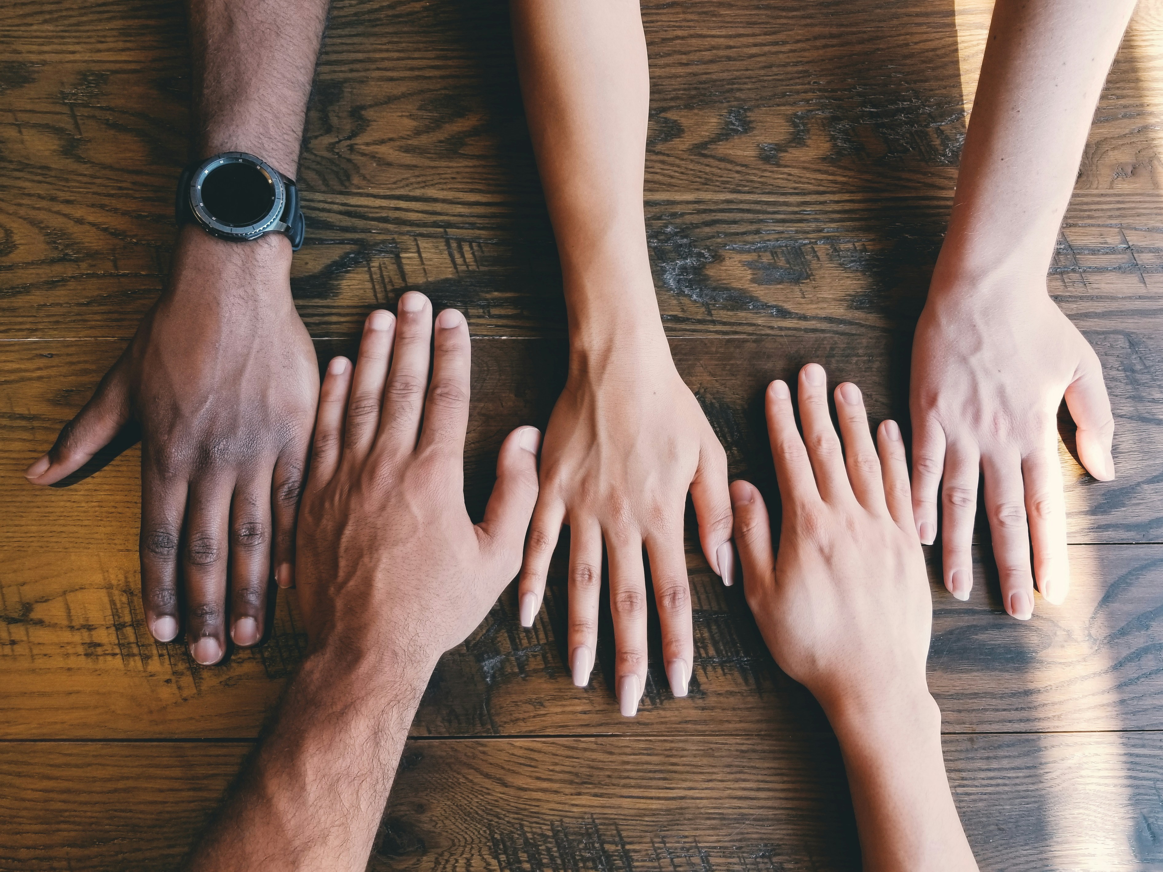 Five hands of different ethnicities and genders side by side on a wood surface. 