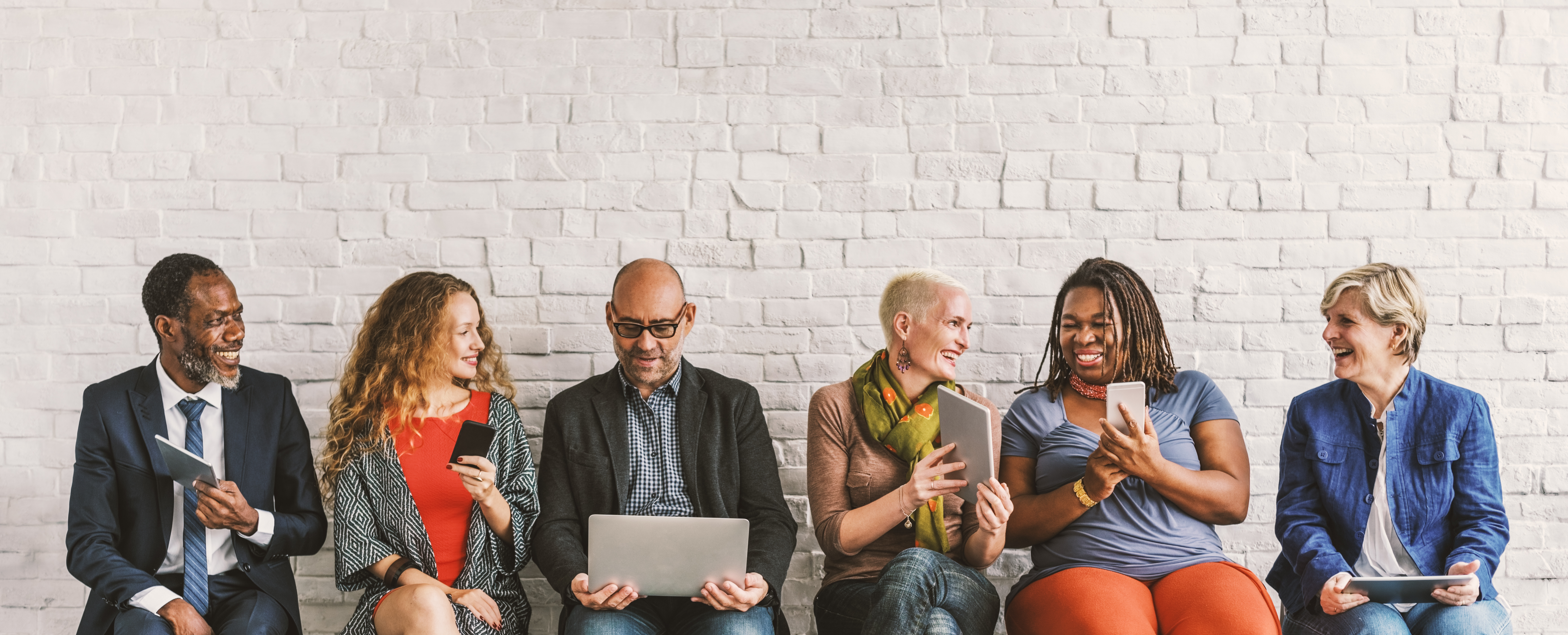 Stock photo of six adults seated in front of a wall, smiling, sharing what is on a phone or laptop