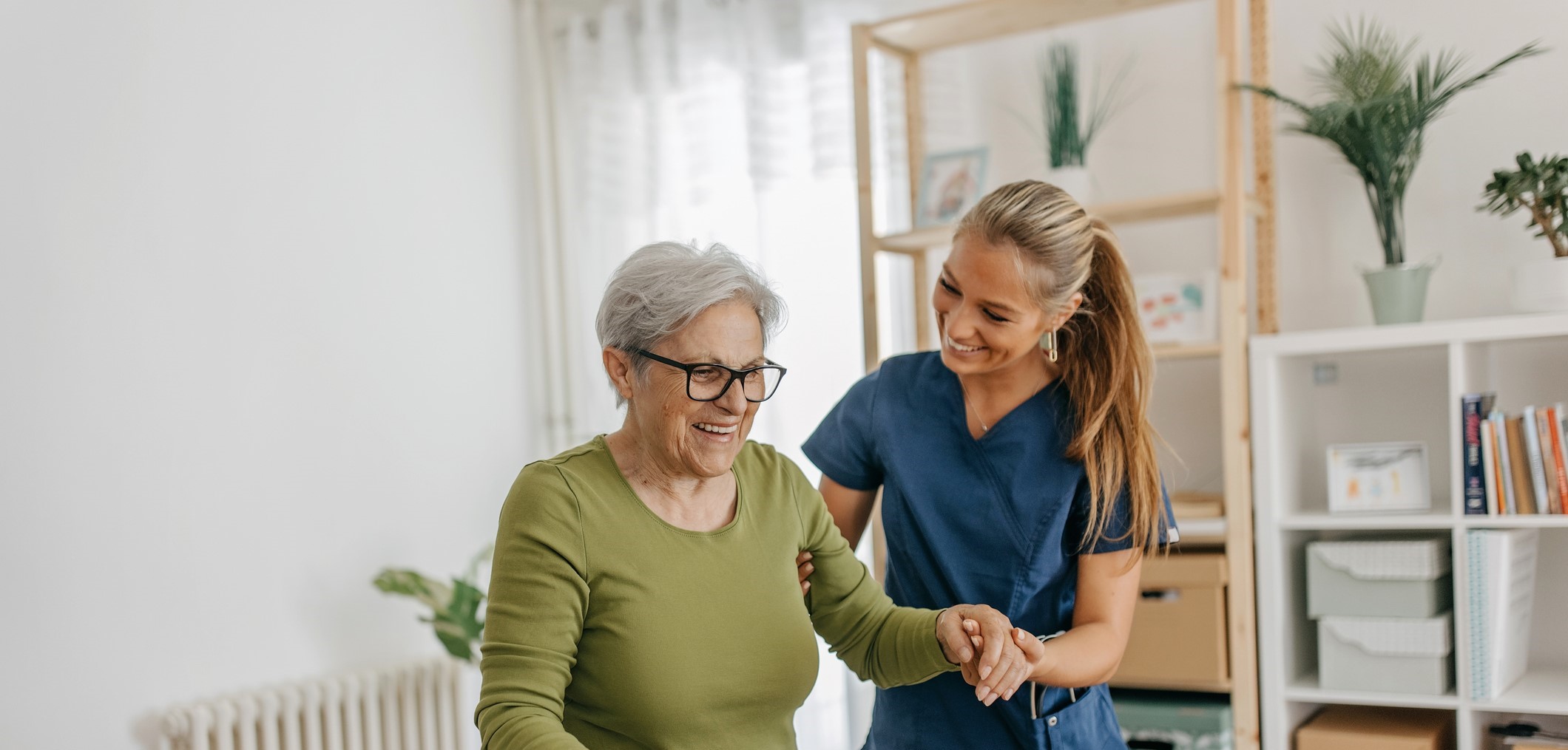 A smiling white elderly woman walking indoors with a cane is assisted by a white female care taker