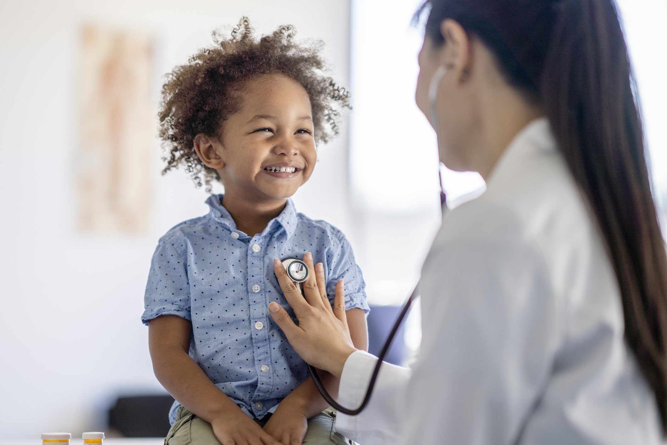 Light brown child sitting on a medical provider table smiling as his doctor listening to his heart with a stethoscope. 