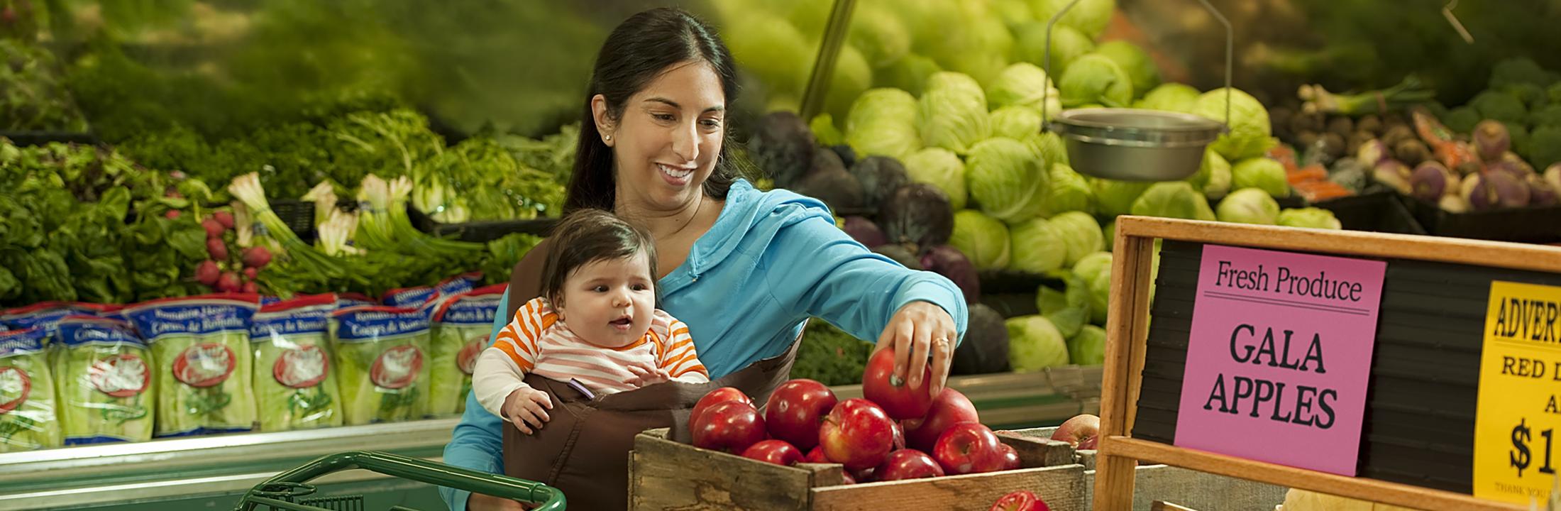 A diverse woman at a supermarket holding an infant while selecting an apple.