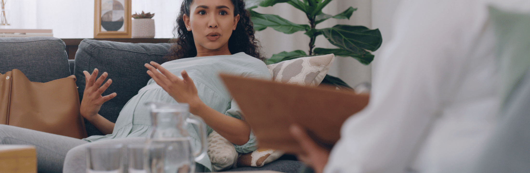 A pregnant woman seated on a plush sofa speaking with a health care provider.