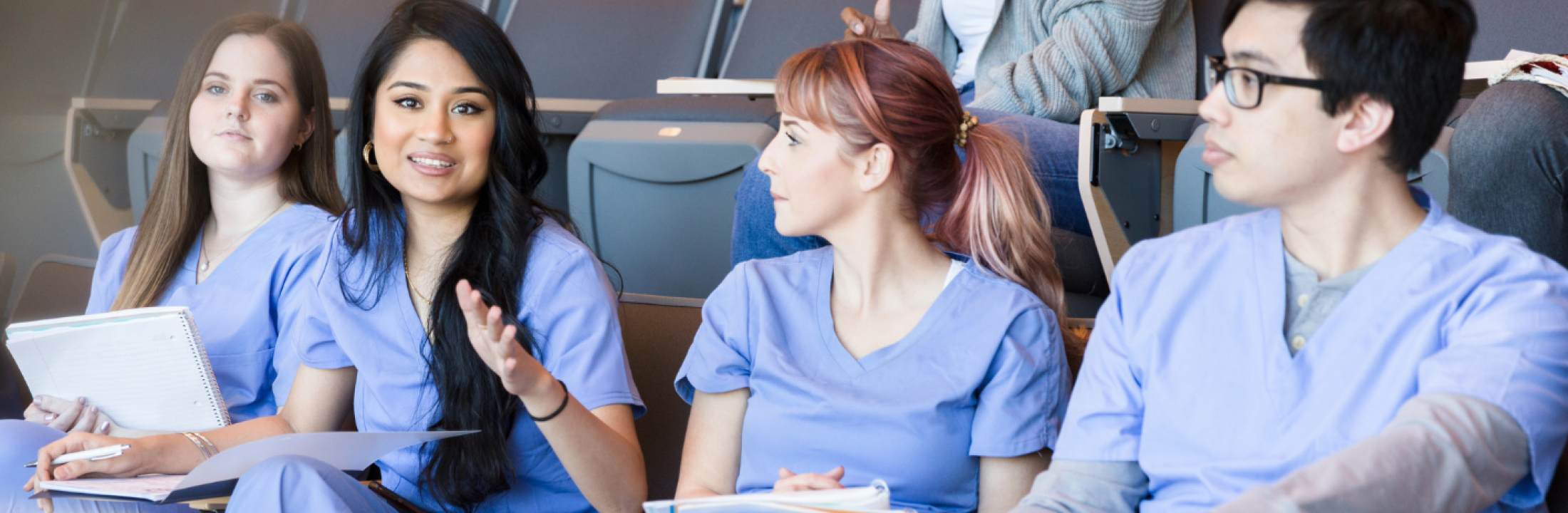 Diverse young adults wearing blue nursing scrubs talk while seated in a college auditorium