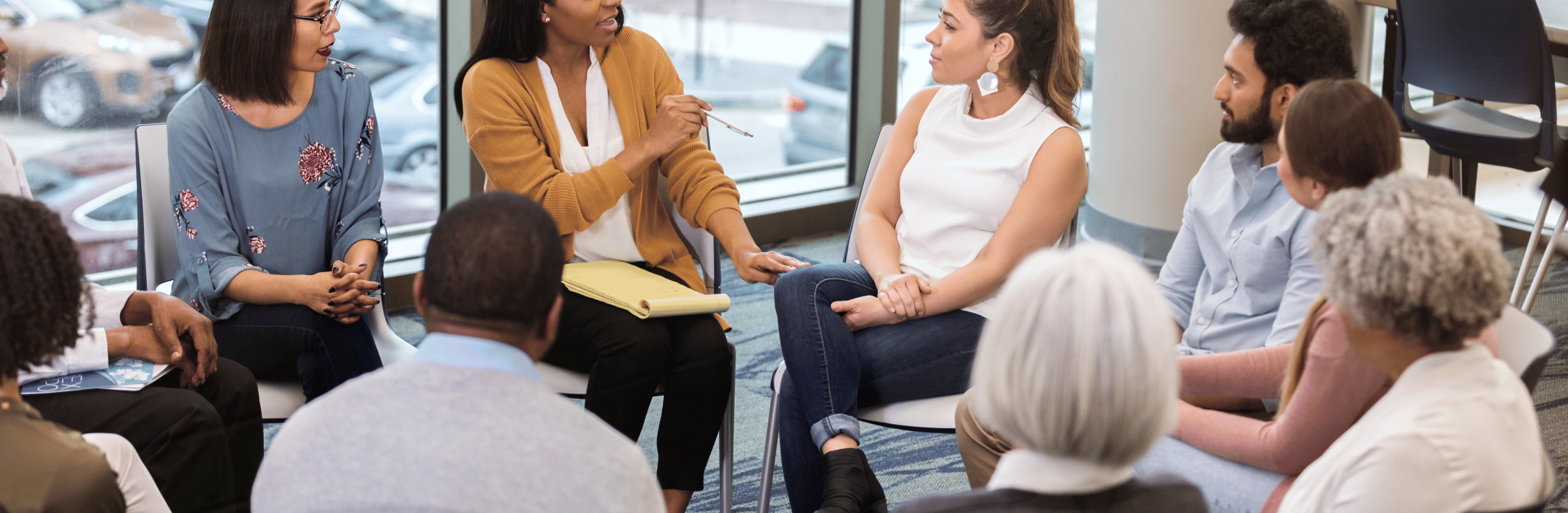 Diverse group of people sitting in a circle with a woman holding a notepad leading the conversation.