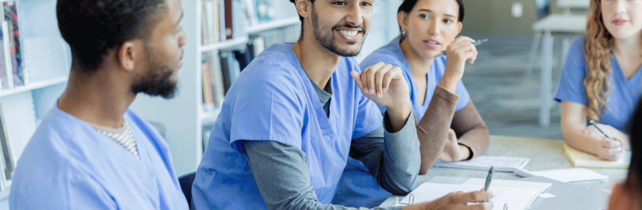 Young adults wearing blue medical scrubs seated in a library talking. 