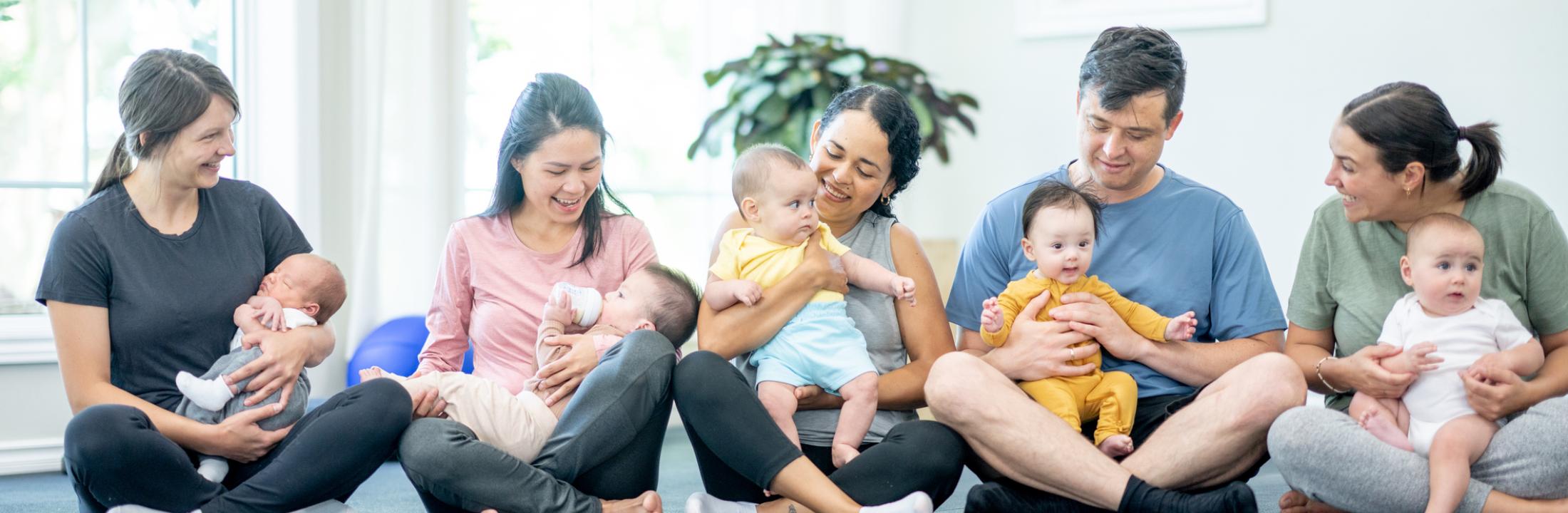 Five parents holding infants as they sit together on a floor.