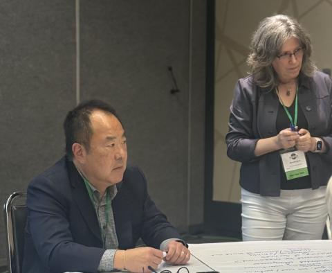 Dr. William Hirota, a man wearing a black suit jacket, seated at a table, and Michelle Davis, a female, wearing a gray suit jacket with white pants, standing, listening to public members at a round table.