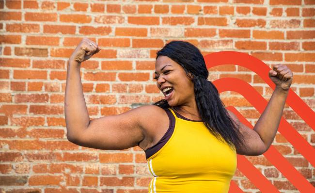 Black woman in a yellow tank top flexing her biceps in front of a brick wall.