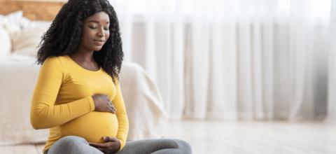 Diverse pregnant woman seated on bedroom floor smiling and meditating.